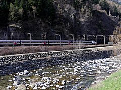 Photographie d'une rivière qui borde la route, avec des galets au premier plan mais des rails au-delà, avec un TGV SNCF qui y passe.
