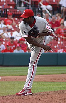 A dark-skinned man with a goatee wearing a gray baseball uniform with red trim and a red baseball cap throws a baseball from a dirt mound