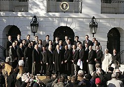 Group of about thirty men wearing suits in front of a white building
