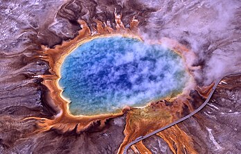 Vue aérienne du Grand Prismatic Spring dans le parc national de Yellowstone entre les États de l’Idaho, du Montana et du Wyoming). De cette source d’origine volcanique très active (une caldeira issue de l’explosion et l’effondrement d’un volcan sur une immense chambre magmatique peu profonde) surgit une eau brûlante, stérile et teintée d’azur (à cause de son acidité), mais ses rives se parent en été de rouge, orange et jaune (par la forte concentration en caroténoïdes antioxydants et photoprotecteurs sécrétés par les algues thermophiles microscopiques qui peuvent s’y développer. Le parc national regroupe à lui seul les deux tiers des geysers de la planète. (définition réelle 1 999 × 1 277)