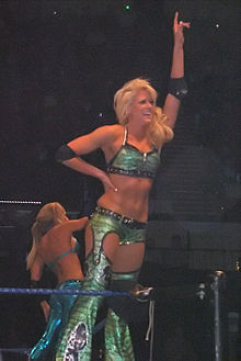 A photo of a blonde Caucasian female, who is posing on the second turnbuckle of a wrestling ring with blue ropes. She has one hand extended into the air, and is pointing upwards with her index finger, while the other hand rests on her waist, and is wearing a green crop top and green trousers. A second blonde woman in visible in the background, wearing a blue crop top and trousers.