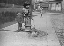 Ein kleines Mädchen lehnt sich kaum tiefer an einen Hydranten und sieht zu, wie sich eine Flasche mit Wasser füllt.