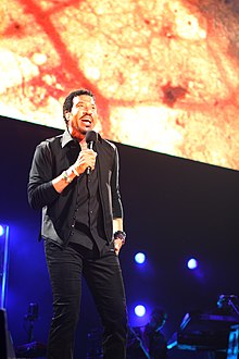 un cantante, maglietta e pantaloni scuri, sul palco con un microfono