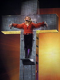 An image showing a blue crystal cross and a woman standing on a platform on the cross. The woman is wearing an orange shirt and dark brown pants. Her hands are spread apart along the cross's breadth to symbolize as if she has been crucified.