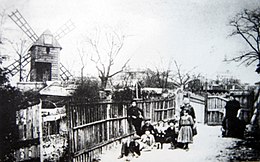 Черно-белое фото нескольких женщин и детей на дорожке между частоколами, хижинами и мельницей позади