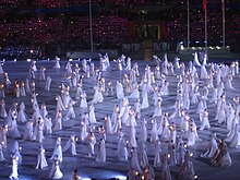 Bir performanstan bir sahnenin fotoğrafı, beyaz tulumlu merkezde birçok dansçı.