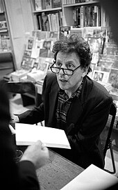 Black and white photograph of a man sitting at a table; he is wearing a suit and glasses, and is being approached by someone carrying an object in their hand.