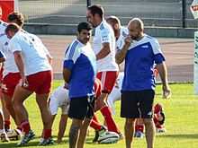 En primer plano, dos hombres de pie en ropa deportiva están charlando, en el fondo, los jugadores de rugby están calentando.