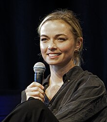 A candid bust photo of a young white woman, wearing a dark blouse; she has blonde hair, is smiling to the camera's left, and is holding a microphone.