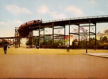 Panorama einer Straßenszene. Freiflächen aus Straßen und einem Park, nur im Bildhintergrund Gebäude. Die obere Bildhälfte wird demoniert von dem technischen Konstrukt der Hochbahntrasse, deren Bauweise aus stählernen Verstrebungen hier fast filigran wirkt. Auf der Hochbahn fährt ein von einer kleinen Dampflok gezogner Wagenzug. In der unteren Bildhälfte sind zum Größenvergleich eine Handvoll Fußgänger und Radfahrer abgebildet.