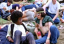 Une photographie couleur montrant des gens du festival de Woodstock de 1969 assis sur l'herbe, au premier plan un dos et un homme blanc se regardent