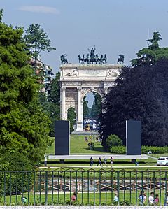 A decorated stone arch seen from a distance through a vista with lawns and trees on the side. A metal fence is in the foregound