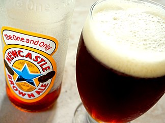 A wide Geordie schooner glass with a stem, filled with dark brown beer with a large foam head. Next to it is a mostly-empty bottle labelled "The One and Only: Newcastle Brown Ale"