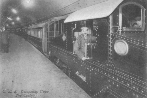 a large electric locomotive sits at a dimly lit platform underground with carriages behind. The cap-wearing driver and his assistant pose for the camera at the controls. Other staff are visible along the platform.