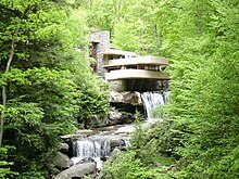 A modernist-style house sits nestled in the woods, with a multilevel terrace hanging over a waterfall