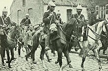 Fotografía en blanco y negro de un pequeño grupo de infantería rodeado de jinetes, portando sables o pistolas.