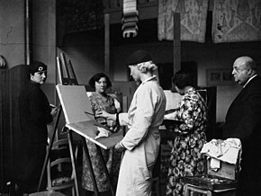 Photo en noir en blanc montrat un homme de profil, chauve, plutôt corpulant, observant plusieurs jeunes femmes artistes, chacune avec son chevalet.