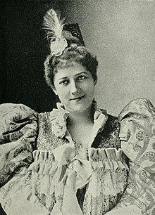 Black and white photograph of Lillian Lewis, wearing a dress with ruffles and large sleeves, and a feathered headpiece