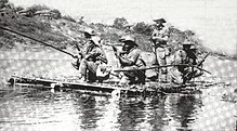 A raft on a river. A man with a rifle stands in the centre, surrounded by other men kneeling on one knee.