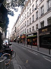 Rue à une voie et contresens cyclable bordée d'immeubles de 5 étages sur la droite, bordée d'arbres minces sur la gauche