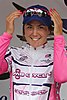 Chloe Hosking on an awards podium after an event. She is a nineteen year old white female with brown hair, blue eyes and a big smile, wearing a pink and white cycling jersey and a blue peaked cap with pink framed mirror sunglasses on top