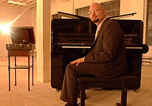 un homme avec un bouc, des lunettes et une boucle d'oreille assis devant un piano droit