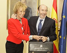 Photographie en couleur d'une femme debout et d'un homme debout à côté d'elle. Tous deux sourient et tiennent le même sac par sa poignée.
