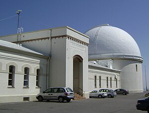 A white one story building with tall thin windows and an alcove entrance; at the right far end, a white domed building is present.