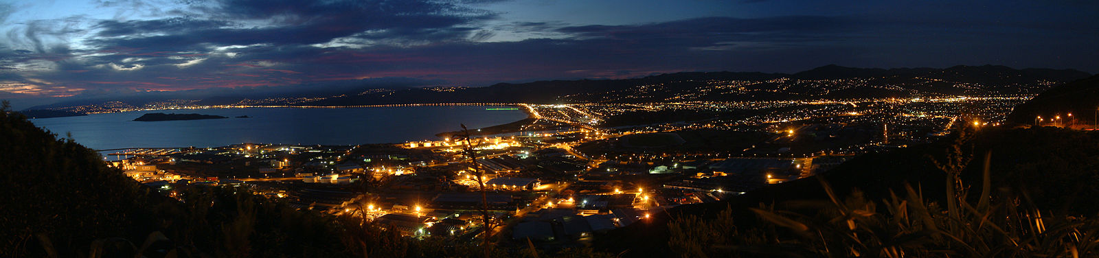 Wellington Harbour and southern Lower Hutt from the top of the Wainuiomata Hill Road (south of the above photo), looking west. Matiu / Somes Island is in the harbour on the left (South), and beyond that the row of lights along State Highway 2, marking the line of the geologic fault, both of which continue up the far side of the valley to the right. The industrial area in the central foreground is Gracefield. In the distance, behind Matiu / Somes Island, are Wellington port and CBD.