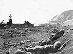 أفراد من الكتيبة الأولى من فوج مشاة البحرية 23 يحفرون في الرمال البركانية على الشاطئ الأصفر رقم 1. يظهر في أعلى اليسار سفينة إنزال على الشاطئ بينما يظهر جبل سوريباتشي أعلى اليمين.