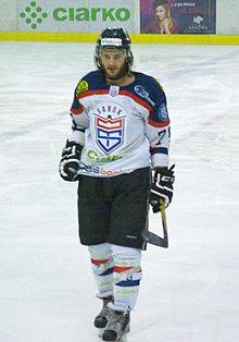 Photographie couleur d’un joueur de hockey sur glace, de face, en pied, portant une barbe de quelques jours et les cheveux dépassant du casque