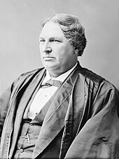 a white man in a suit, facing left-front, wearing judicial robes