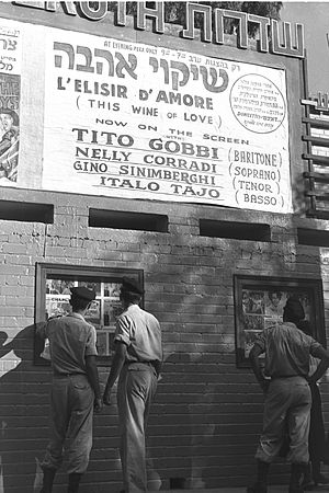 חיילים במדים, כומתות על ראשם צופים בכרזות בכניסה לקולנוע שדרות, בשנת 1955. מתחת לשלט שם בית הקולנוע כרזה לסרט "שיקוי האהבה"