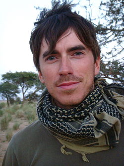 Portrait photograph of Simon Reeve; a white male. A BBC publicity photo, taken on location during his travels for the 'Tropic of Cancer' television series. Showing his head to his chest to just below shoulder level, Reeve, looking straight at the camera, has an engaging but neutral look on his face. Mouth closed, eyes blue-green topped with thick dark brown eyebrows, head covered in slightly unkempt dark brown hair, his face and top lip covered in very short ginger coloured stubble. He wears a green T-shirt, with an Arabic style head scarf loosely wrapped around his neck.