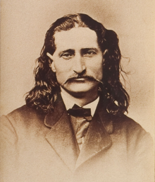 A slightly smiling man dressed in an overcoat and sporting a mustache and shoulder-length, curly hair stares ahead.
