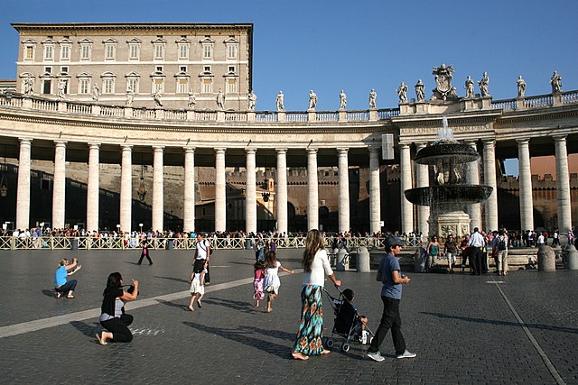 Farbfotografie von einem Platz mit Säulengang und Figuren darauf. Ein Springbrunnen ist auf der rechten Seite zu sehen und einige Menschen sind im Vordergrund. Hinter einer Absperrung am Säulengang hat sich eine lange Menschenschlange gesammelt. Über dem Säulengang ist links der obere Bereich eines Gebäudes mit vielen Fenstern zu sehen.