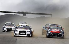 Cinco carros esportivos vistos de frente em uma pista de asfalto e levantando uma nuvem de poeira.