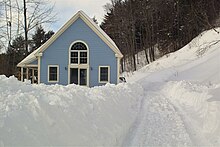 Ein hellbaues Haus steht auf einem Hang inmitten tiefen Schnees. Am rechen unteren Bildrand sind Räderspuren erkennbar.