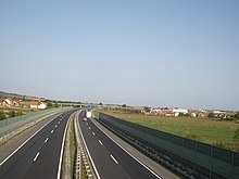 Autostradă cu șase benzi trecând printr-o zonă rurală, separată prin panouri antizgomot.