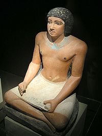Крашеная, реалистичная, каменная статуя черноволосого мужчины, сидящего со скрещёнными ногами и держащего каменное изображение папирусного свитка на коленях