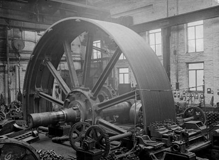Flywheel for a large textile mill engine 1900, set up to machine grooves for the rope drives simultaneously. The saddle with two tool posts to the front. The wheel is rotated by two pinions driving via the cast-in barring gear teeth in the flywheel rim. Temporary wedges are securing the spokes to the hub of the wheel. A travelling crane behind and above.[63][98]