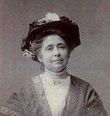 An older white woman, wearing large black hat, a high-collared white lace blouse or dress. Her greying hair is in a bouffant updo.
