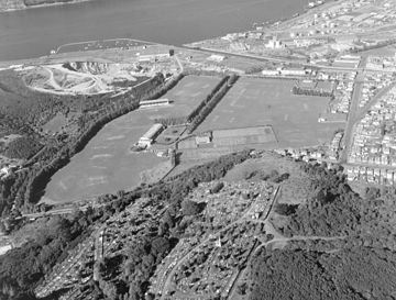 An aerial view in 1955, looking south. The Caledonian Ground had not been built, but the University Oval, former Art Gallery (centre left) and former Aquarium (centre) are clearly visible. Logan Point Quarry and the Otago Harbour are visible in the background. Logan Park, Dunedin (1955).jpg