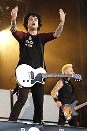 Два члена рок-группы Green Day показаны на сцене во время концерта. Слева направо певец / гитарист Билли Джо Армстронг и бас-гитарист Майк Дирнт. Позади них ряд больших кабинетов гитарных динамиков. Билли Джо обеими руками показывает на публику. 