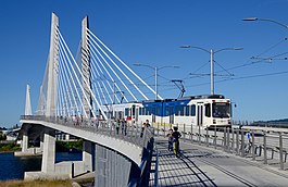 The Tilikum Crossing bridge with a MAX train traveling northbound and pedestrians walking alongside