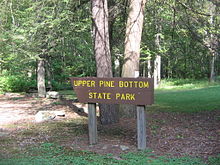 A brown wooden sign has Upper Pine Bottom State Parkt in yellow letters, with large tree trunks, rocks, and a picnic table in the background