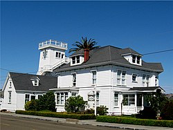 A Victorian building with a Mansard roof, three stories tall, painted white with a taller water tower wrapped by a staircase behind it