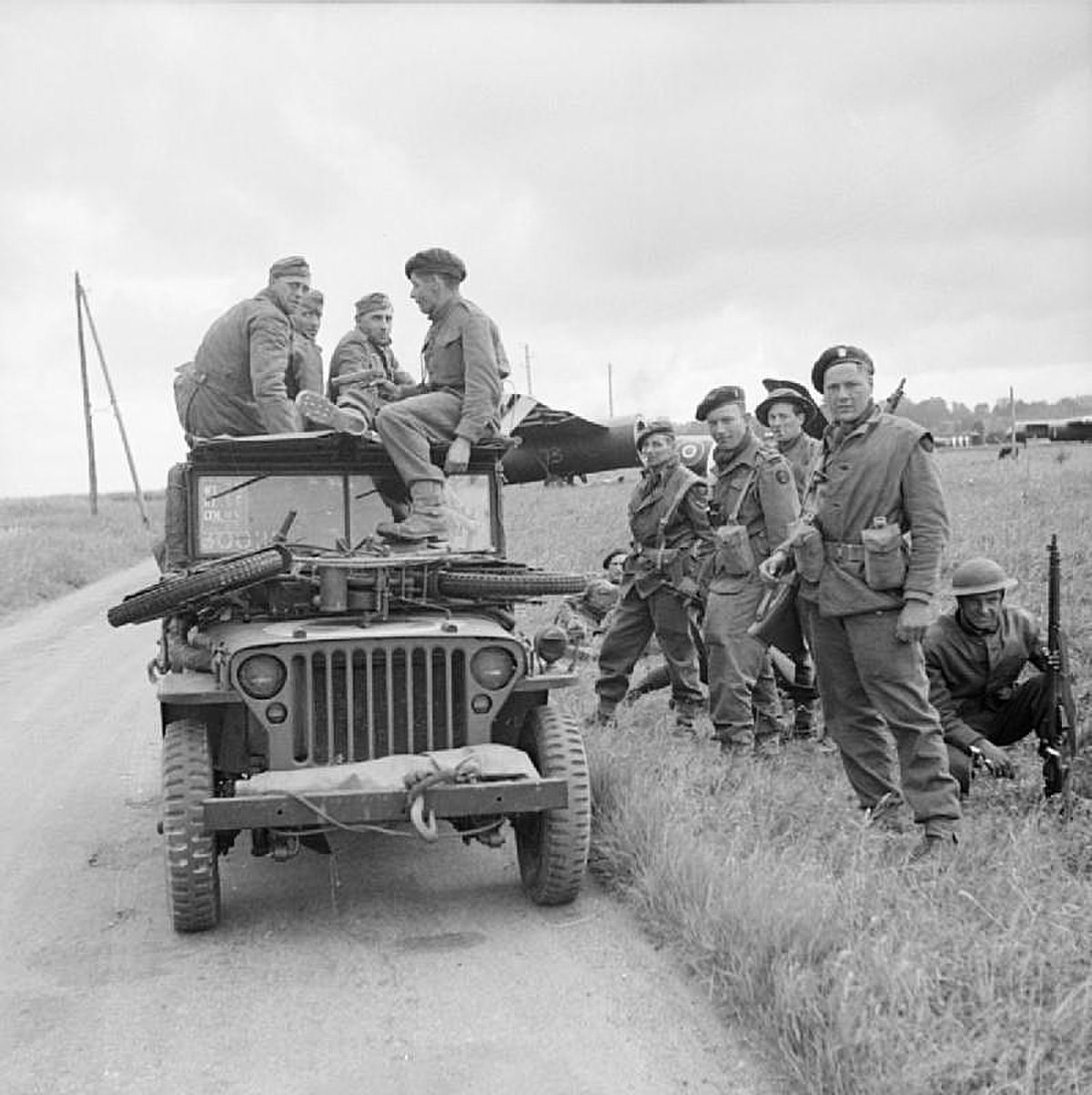 A black-and-white photograph of a jeep with soldiers sitting on top and standing beside it. The soldiers sitting on the jeep are three German soldiers and one British soldier who is interrogating the Germans. On the bonnet of the jeep is small motorcycle， while in the background is a Horsa glider