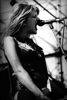 photo en noir et blanc d'une femme criant dans un micro tout en jouant de la guitare.
