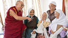 The 14th Dalai Lama with elderly Tibetan Muslims. The Dalai Lama is wearing gold and red Buddhist robes, while the Muslims are dressed in traditional Tibetan casual wear. A Muslim man sports a white taqiyah cap, while the Muslim women wear white headscarves.